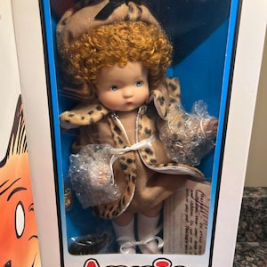 May include: A collector's doll named Annie, dressed in a brown and black leopard print coat with a brown hat. The doll has curly red hair and blue eyes. The doll is packaged in a white box with blue trim and the text "Annie Collector Doll".