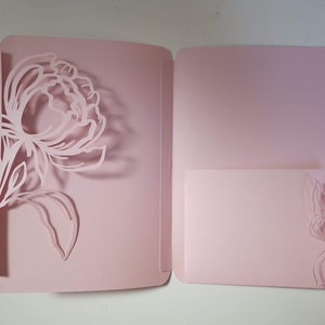 Puede incluir: Carpeta de bolsillo de papel rosa claro con un diseño floral troquelado. La carpeta abierta revela un bolsillo más pequeño en el lado derecho, también con un acento floral. El diseño es una delicada peonía.