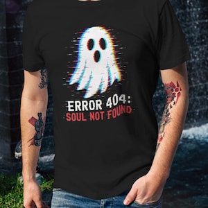 May include: A black short-sleeved t-shirt featuring a white ghost graphic with a digital glitch effect. Below the ghost, the text reads ERROR 404: SOUL NOT FOUND in white and red lettering. The shirt is paired with blue denim jeans.