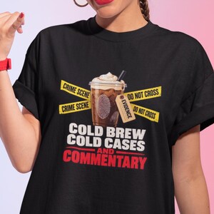 May include: A black t-shirt featuring a graphic of an iced coffee drink surrounded by yellow crime scene tape. The text reads COLD BREW, COLD CASES, AND COMMENTARY. The design includes an evidence tag and the phrases CRIME SCENE and DO NOT CROSS.