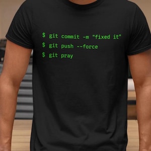 Puede incluir: Una camiseta negra de manga corta con texto verde que dice: $ git commit -m "fixed it", $ git push --force y $ git pray. La prenda tiene un cuello redondo y un corte estándar, mostrando comandos de programación en estilo terminal.