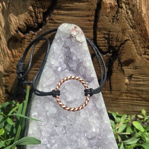 May include: A bracelet featuring a circular, twisted copper-colored metal ring centered on a thin black cord. The cord is adjustable with sliding knots on each side of the metal ring, creating a simple design that secures around the wrist.