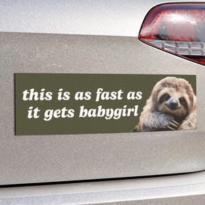 May include: A rectangular bumper sticker with the text "this is as fast as it gets babygirl" in white font on an olive green background. A smiling sloth is on the right side of the sticker.