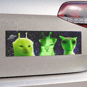 May include: A bumper sticker with a green alien cat design. The sticker features three cats with alien antennas and glowing green eyes. The background is a dark blue with stars.