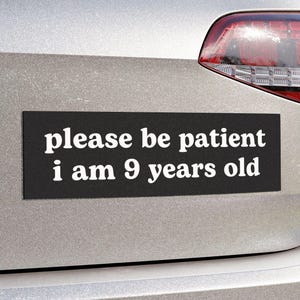 Könnte beinhalten: Ein schwarzer, rechteckiger Autoaufkleber mit weißem Text, der "please be patient i am 9 years old" lautet, ist am Heck eines silbernen Autos angebracht. Das Rücklicht des Autos ist sichtbar.