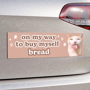 Puede incluir: Una pegatina para parachoques de coche rosa y blanca con un gato con la lengua fuera. La pegatina dice "On my way to buy myself bread" con estrellas alrededor del texto.