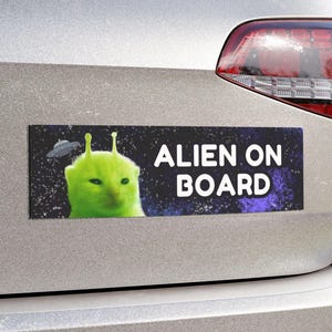 Op de afbeelding: Een zilveren auto met een bumper sticker waarop staat "Alien on Board". De sticker toont een groene alien met grote ogen op een zwarte achtergrond met sterren.