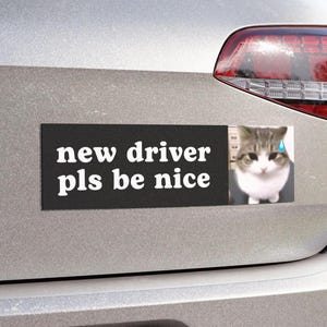 May include: A rectangular bumper sticker with a black background and white text that reads new driver pls be nice. To the right of the text is a photographic image of a grey and white tabby cat with a blue teardrop icon near its eye.