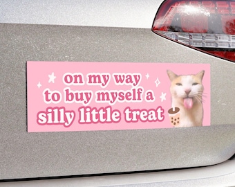 May include: Pink car bumper sticker with a white cat with a pink tongue sticking out. The sticker says "on my way to buy myself a silly little treat" with stars and sparkles around the text.