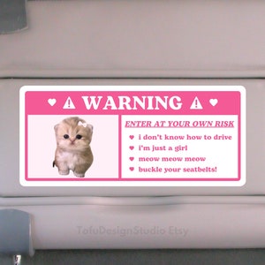 May include: A pink and white car visor sticker with a kitten graphic and the text "A WARNING" and "ENTER AT YOUR OWN RISK". The text includes "I don't know how to drive", "I'm just a girl", "meow meow meow", and "buckle your seatbelts!"