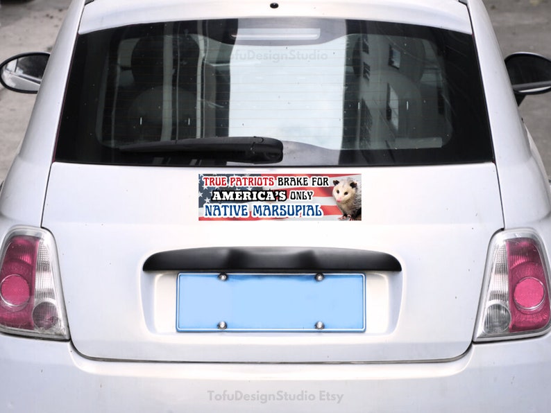 Puede incluir: Un coche blanco con una pegatina en el parachoques que dice "True Patriots Brake for America's Only Native Marsupial". La pegatina presenta una bandera estadounidense y una imagen de una zarig&uuml;eya.