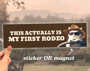 Könnte beinhalten: Ein brauner rechteckiger Aufkleber oder Magnet mit dem Text "This actually is my first rodeo" und einer weißen Katze mit Sonnenbrille und Cowboyhut, die ein Auto fährt.