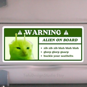 Op de afbeelding: Een groen waarschuwingsbord voor een autozonnescherm met een groene alienkat met antennes en de tekst "A WARNING A ALIEN ON BOARD" en een lijst met instructies: "zib zib zib blah blah blah", "glorp glorp gnarp" en "doe uw gordels om".