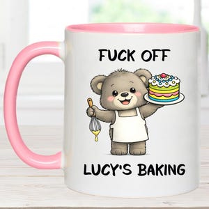 May include: White ceramic mug with a pink handle and interior. The mug features the text "FUCK OFF" above an illustration of a teddy bear holding a cake and whisk. The text "LUCY'S BAKING" is below the bear.