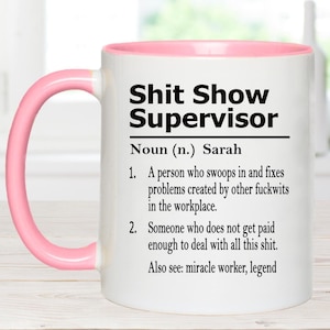 May include: A white ceramic mug with a pink handle and interior rim. The front features black text reading: Shit Show Supervisor, Noun (n.) Sarah, 1. A person who swoops in and fixes problems created by other fuckwits in the workplace. 2. Someone who does not get paid enough to deal with all this shit. Also see: miracle worker, legend.
