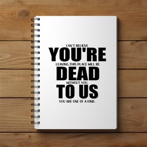 May include: A white spiral-bound notebook with black text that reads "CAN'T BELIEVE YOU'RE LEAVING. THIS PLACE WILL BE DEAD WITHOUT YOU. TO US. YOU ARE ONE OF A KIND." The notebook is on a wooden surface.