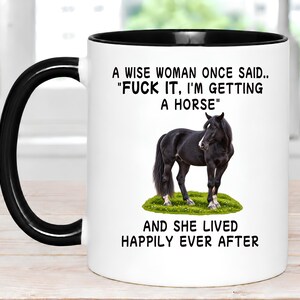 May include: A white ceramic mug with a black handle and rim, featuring a black horse standing on a patch of green grass. The text reads: A WISE WOMAN ONCE SAID.. "FUCK IT, I'M GETTING A HORSE" AND SHE LIVED HAPPILY EVER AFTER in black lettering.