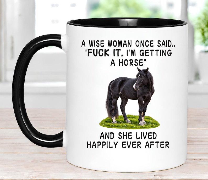 May include: A white ceramic mug with a black handle and rim, featuring a black horse standing on a patch of green grass. The text reads: A WISE WOMAN ONCE SAID.. "FUCK IT, I'M GETTING A HORSE" AND SHE LIVED HAPPILY EVER AFTER in black lettering.