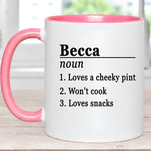 May include: A white ceramic mug with a pink handle and interior rim. The front features black text that reads: Becca noun 1. Loves a cheeky pint 2. Won't cook 3. Loves snacks. The design is printed in a dictionary definition style with a horizontal line.