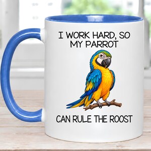 May include: A white ceramic mug with a blue interior and handle. The front features a colorful illustration of a blue and yellow parrot perched on a branch, with the text "I WORK HARD, SO MY PARROT CAN RULE THE ROOST" printed in black.