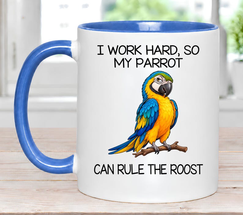 May include: A white ceramic mug with a blue interior and handle. The front features a colorful illustration of a blue and yellow parrot perched on a branch, with the text "I WORK HARD, SO MY PARROT CAN RULE THE ROOST" printed in black.