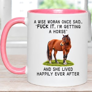 May include: A white ceramic mug with a pink interior and handle. The front features a brown horse standing on green grass with the text: A WISE WOMAN ONCE SAID.. "FUCK IT, I'M GETTING A HORSE" AND SHE LIVED HAPPILY EVER AFTER in black lettering.