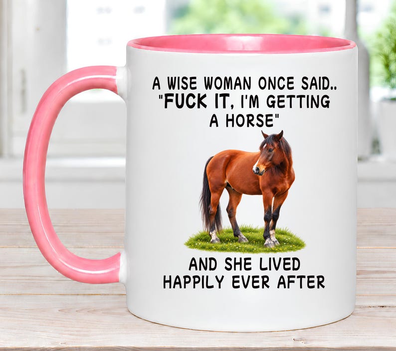 May include: A white ceramic mug with a pink interior and handle. The front features a brown horse standing on green grass with the text: A WISE WOMAN ONCE SAID.. "FUCK IT, I'M GETTING A HORSE" AND SHE LIVED HAPPILY EVER AFTER in black lettering.