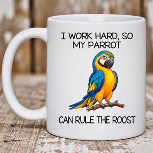 May include: A white ceramic mug featuring a colorful illustration of a blue and yellow macaw parrot perched on a brown branch. The text printed on the mug reads: I WORK HARD, SO MY PARROT CAN RULE THE ROOST. The mug has a curved handle on the left side.
