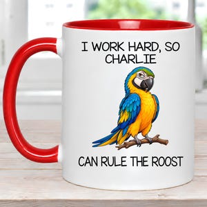 May include: A white ceramic mug with a bright red handle and interior rim. The front features a colorful blue and yellow macaw perched on a branch, with the black text "I WORK HARD, SO CHARLIE CAN RULE THE ROOST" printed above and below the bird.