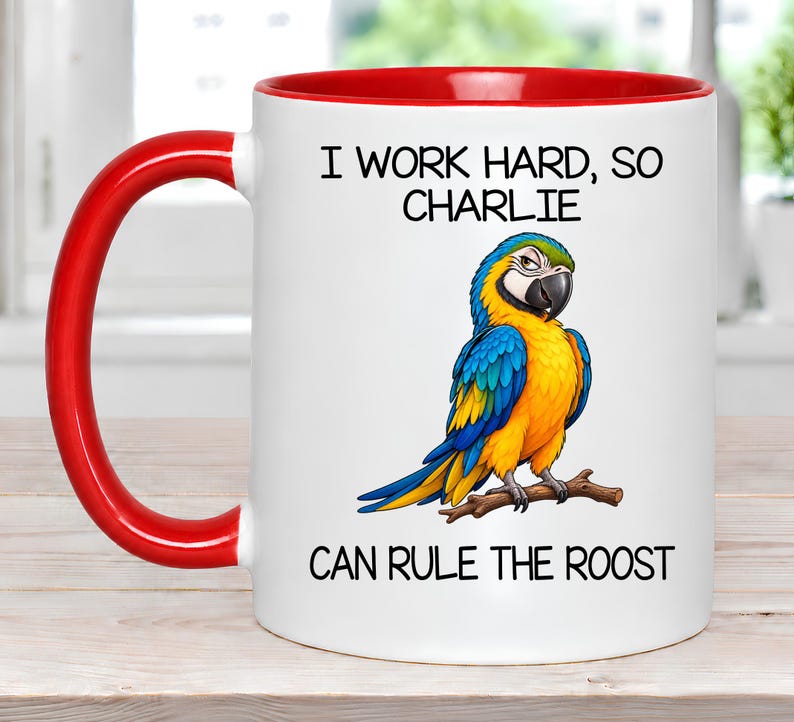 May include: A white ceramic mug with a bright red handle and interior rim. The front features a colorful blue and yellow macaw perched on a branch, with the black text "I WORK HARD, SO CHARLIE CAN RULE THE ROOST" printed above and below the bird.