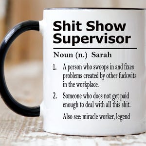 May include: A white ceramic mug with a black handle and interior rim. The front features black text that reads: Shit Show Supervisor, Noun (n.) Sarah, 1. A person who swoops in and fixes problems created by other fuckwits in the workplace. 2. Someone who does not get paid enough to deal with all this shit. Also see: miracle worker, legend.