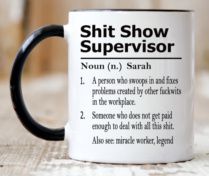May include: A white ceramic mug with a black handle and interior rim. The front features black text that reads: Shit Show Supervisor, Noun (n.) Sarah, 1. A person who swoops in and fixes problems created by other fuckwits in the workplace. 2. Someone who does not get paid enough to deal with all this shit. Also see: miracle worker, legend.