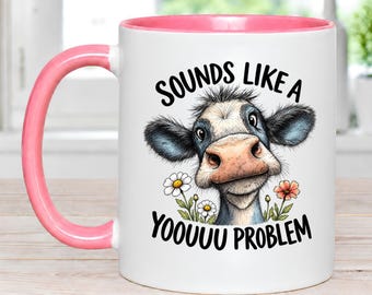 May include: A white ceramic mug with a pink handle and interior rim. The front features a graphic of a cow with black and white fur, surrounded by small daisies and orange flowers, with the black text "SOUNDS LIKE A YOOOUU PROBLEM" printed above and below.
