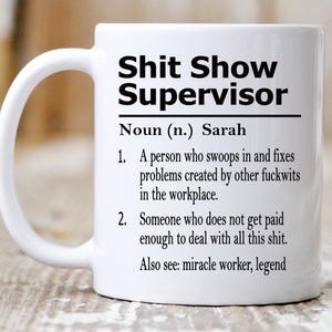 May include: A white ceramic mug featuring black printed text that reads: Shit Show Supervisor, Noun (n.) Sarah, 1. A person who swoops in and fixes problems created by other fuckwits in the workplace. 2. Someone who does not get paid enough to deal with all this shit. Also see: miracle worker, legend.