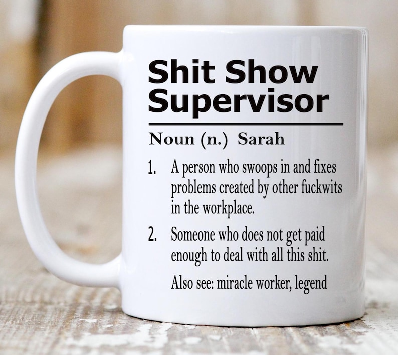 May include: A white ceramic mug featuring black printed text that reads: Shit Show Supervisor, Noun (n.) Sarah, 1. A person who swoops in and fixes problems created by other fuckwits in the workplace. 2. Someone who does not get paid enough to deal with all this shit. Also see: miracle worker, legend.