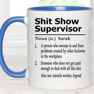 May include: A white ceramic mug with a blue handle and interior rim. The front features black text that reads: Shit Show Supervisor, Noun (n.) Sarah, 1. A person who swoops in and fixes problems created by other fuckwits in the workplace. 2. Someone who does not get paid enough to deal with all this shit. Also see: miracle worker, legend.