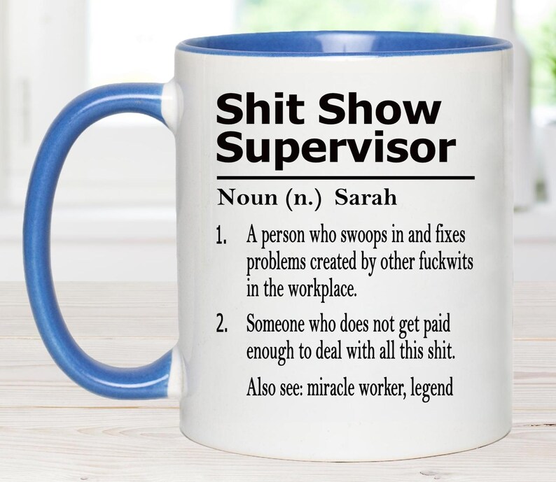 May include: A white ceramic mug with a blue handle and interior rim. The front features black text that reads: Shit Show Supervisor, Noun (n.) Sarah, 1. A person who swoops in and fixes problems created by other fuckwits in the workplace. 2. Someone who does not get paid enough to deal with all this shit. Also see: miracle worker, legend.