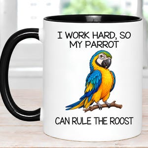 May include: A white ceramic mug with a black handle and rim. The front features a colorful illustration of a blue and yellow macaw perched on a brown branch. The text reads: I WORK HARD, SO MY PARROT CAN RULE THE ROOST in black lettering.