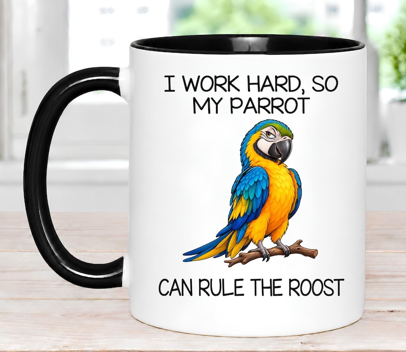 May include: A white ceramic mug with a black handle and rim. The front features a colorful illustration of a blue and yellow macaw perched on a brown branch. The text reads: I WORK HARD, SO MY PARROT CAN RULE THE ROOST in black lettering.