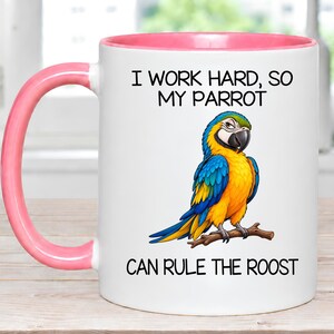 May include: A white ceramic mug with a pink handle and interior rim. The front features a colorful illustration of a blue and yellow macaw perched on a branch, with the black text: I WORK HARD, SO MY PARROT CAN RULE THE ROOST.