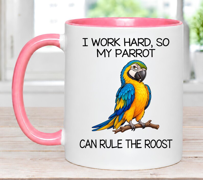 May include: A white ceramic mug with a pink handle and interior rim. The front features a colorful illustration of a blue and yellow macaw perched on a branch, with the black text: I WORK HARD, SO MY PARROT CAN RULE THE ROOST.
