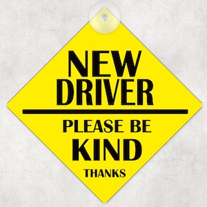 May include: Yellow diamond-shaped sign with black text that reads "NEW DRIVER PLEASE BE KIND THANKS".