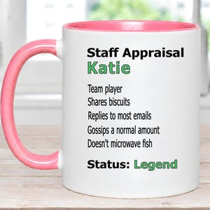 May include: A white ceramic mug with a pink handle and interior rim. The text reads: Staff Appraisal Katie, Team player, Shares biscuits, Replies to most emails, Gossips a normal amount, Doesn't microwave fish, Status: Legend. The name Katie and the word Legend are in green.