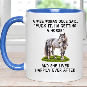 May include: A white ceramic mug with a blue interior and handle. The front features a dapple gray horse standing on green grass, with the black text: A WISE WOMAN ONCE SAID.. "FUCK IT, I'M GETTING A HORSE" AND SHE LIVED HAPPILY EVER AFTER.