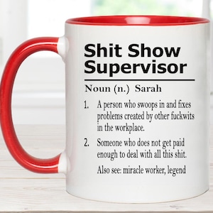 May include: A white ceramic mug with a red handle and interior rim. The front features black text reading: Shit Show Supervisor, Noun (n.) Sarah, 1. A person who swoops in and fixes problems created by other fuckwits in the workplace. 2. Someone who does not get paid enough to deal with all this shit. Also see: miracle worker, legend.