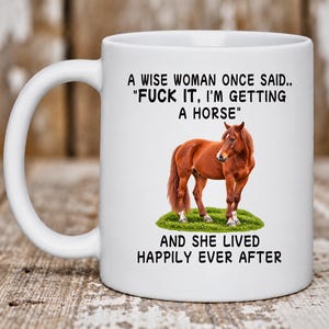 May include: A white ceramic mug featuring a brown horse standing on a patch of green grass. The text reads: A WISE WOMAN ONCE SAID.. "FUCK IT, I'M GETTING A HORSE" AND SHE LIVED HAPPILY EVER AFTER. The mug has a curved handle and a smooth finish.