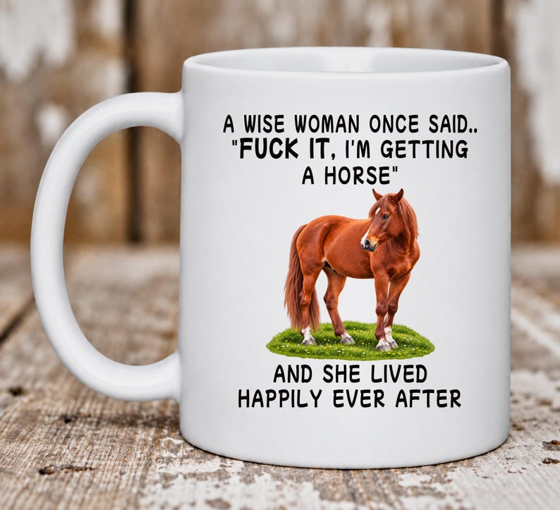 May include: A white ceramic mug featuring a brown horse standing on a patch of green grass. The text reads: A WISE WOMAN ONCE SAID.. "FUCK IT, I'M GETTING A HORSE" AND SHE LIVED HAPPILY EVER AFTER. The mug has a curved handle and a smooth finish.