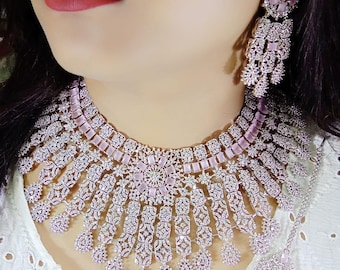 May include: A light pink and silver necklace and earring set. The necklace is a bib style, and the earrings are long and dangling. The jewellery features intricate detailing and sparkling embellishments. The set is designed to be worn together.