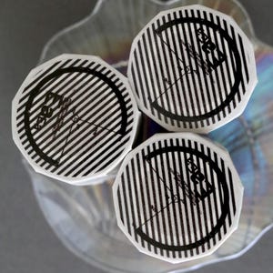 May include: Overhead shot of three rolls of washi tape. Each roll has a white background with black vertical stripes and a black circular design with text. The rolls are on a clear, decorative glass dish.