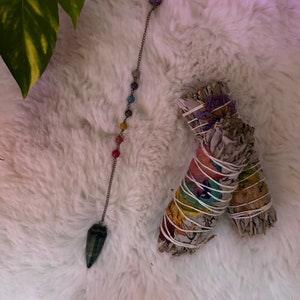 May include: A pendulum with a dark green crystal point and a chain with colorful beads, alongside two bundles of white sage wrapped with string and colorful paper. The items are arranged on a fluffy white surface.